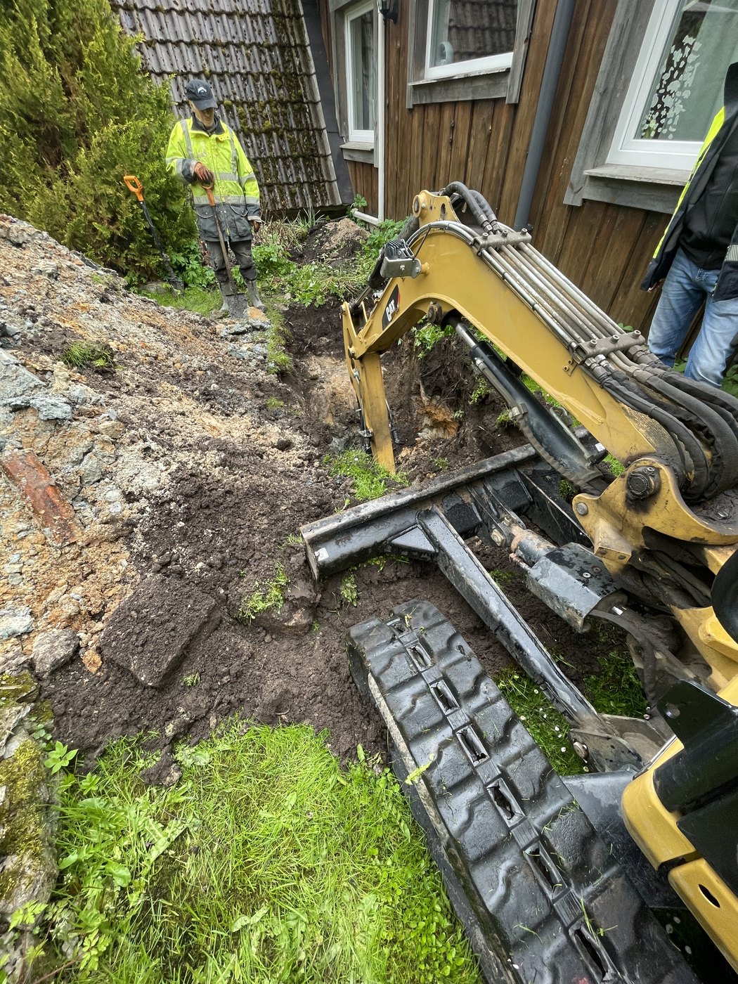 Åpen dreneringsgrøft langs husvegg med gravemaskin og to arbeidere